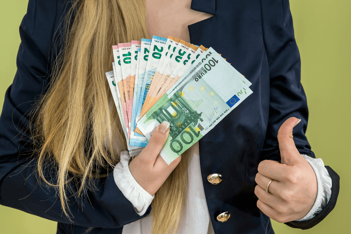 Mulher loira vestida de forma profissional segurando um leque de notas de Euro e fazendo sinal de positivo com a mão, simbolizando os resultados financeiros de quem tem o perfil correto para trabalhar no Superpay.me.