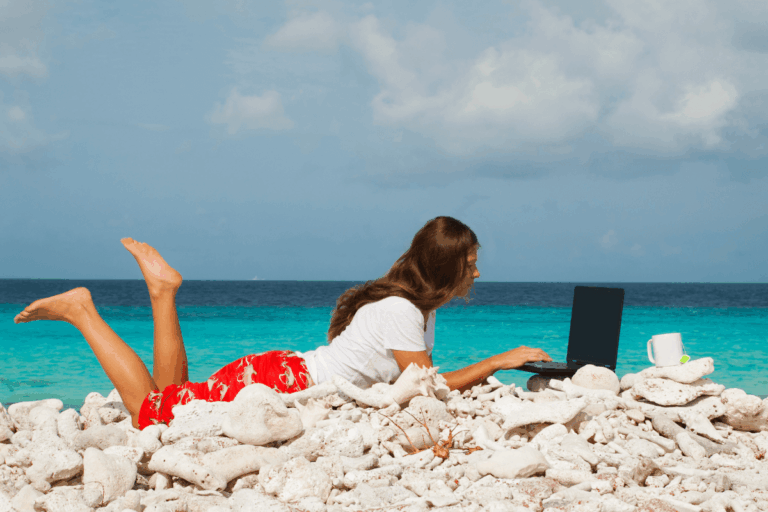 Mulher deitada de bruços em uma praia de pedras brancas trabalhando no notebook com o mar turquesa ao fundo, ilustrando o imaginário popular sobre quais as vantagens e desvantagens de ser um nômade digital.