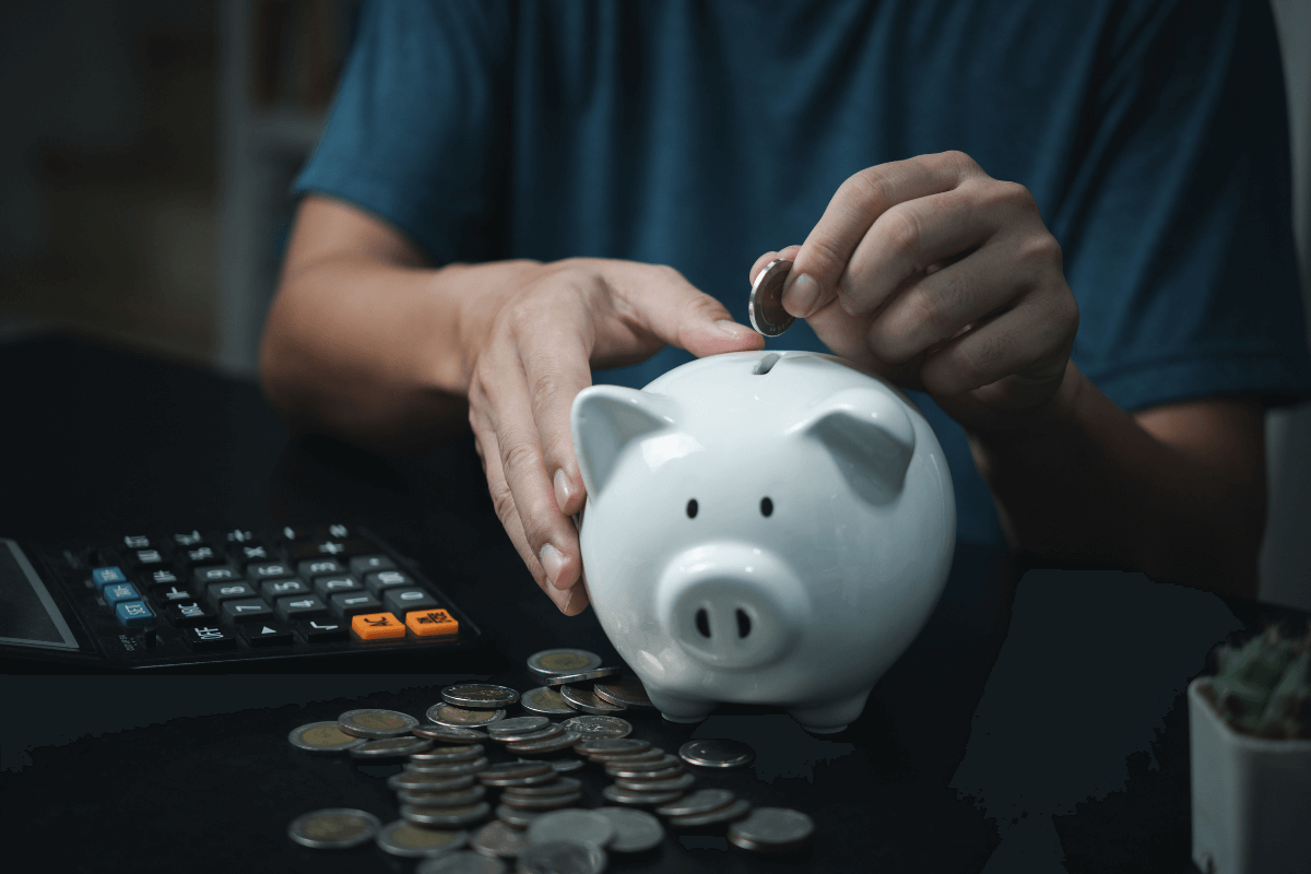 Pessoa depositando moeda em um cofrinho branco ao lado de uma calculadora, ilustrando o acúmulo e a conversão de pontos do Microsoft Rewards em dinheiro real.