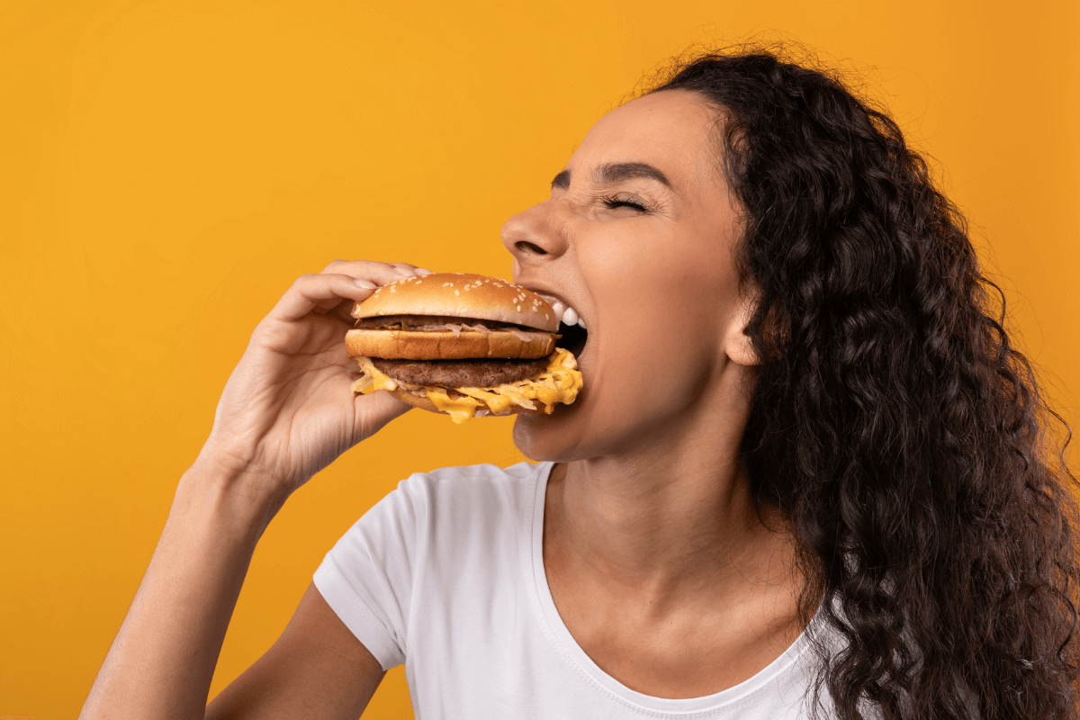 Mulher feliz mordendo um hambúrguer em fundo amarelo, ilustrando a satisfação de trocar pontos do Microsoft Rewards por vouchers de comida no iFood ou Uber Eats.