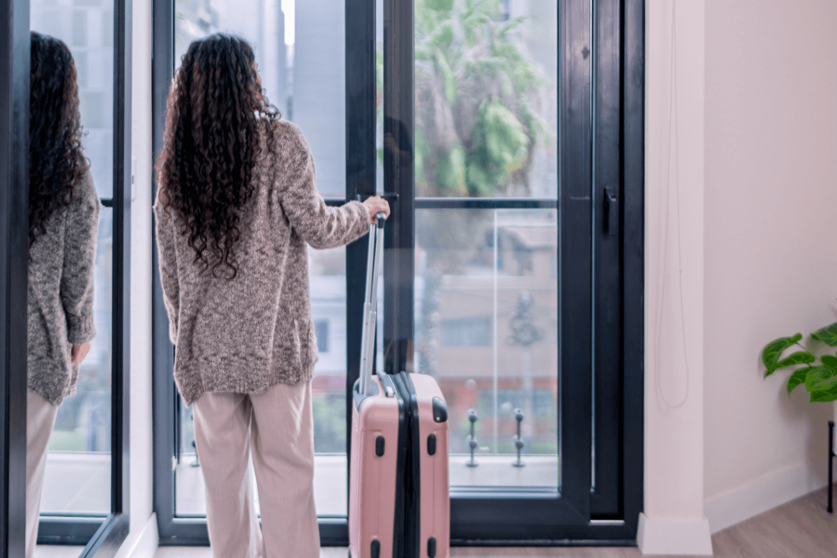 A solidão e a transitoriedade das relações: o peso emocional das despedidas constantes. Mulher de cabelos cacheados, vestindo roupas confortáveis, parada de costas segurando uma mala rosa diante de uma grande porta de vidro, ilustrando o momento de partida e a solidão recorrente na vida de um nômade digital.
