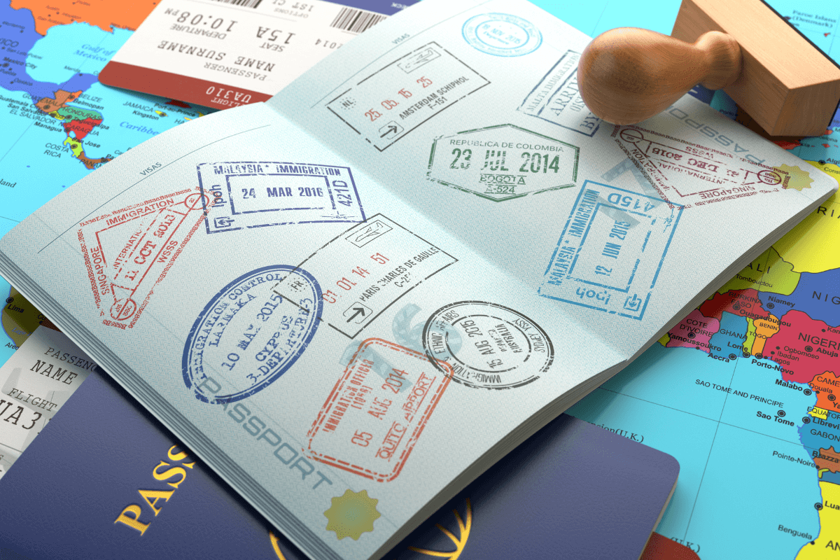 Burocracia de vistos e residência fiscal: o desafio dos carimbos e leis de imigração. Passaporte aberto repleto de carimbos de imigração variados e um carimbo de madeira sobre um mapa mundi colorido, ilustrando a complexidade da burocracia de vistos para nômades digitais.