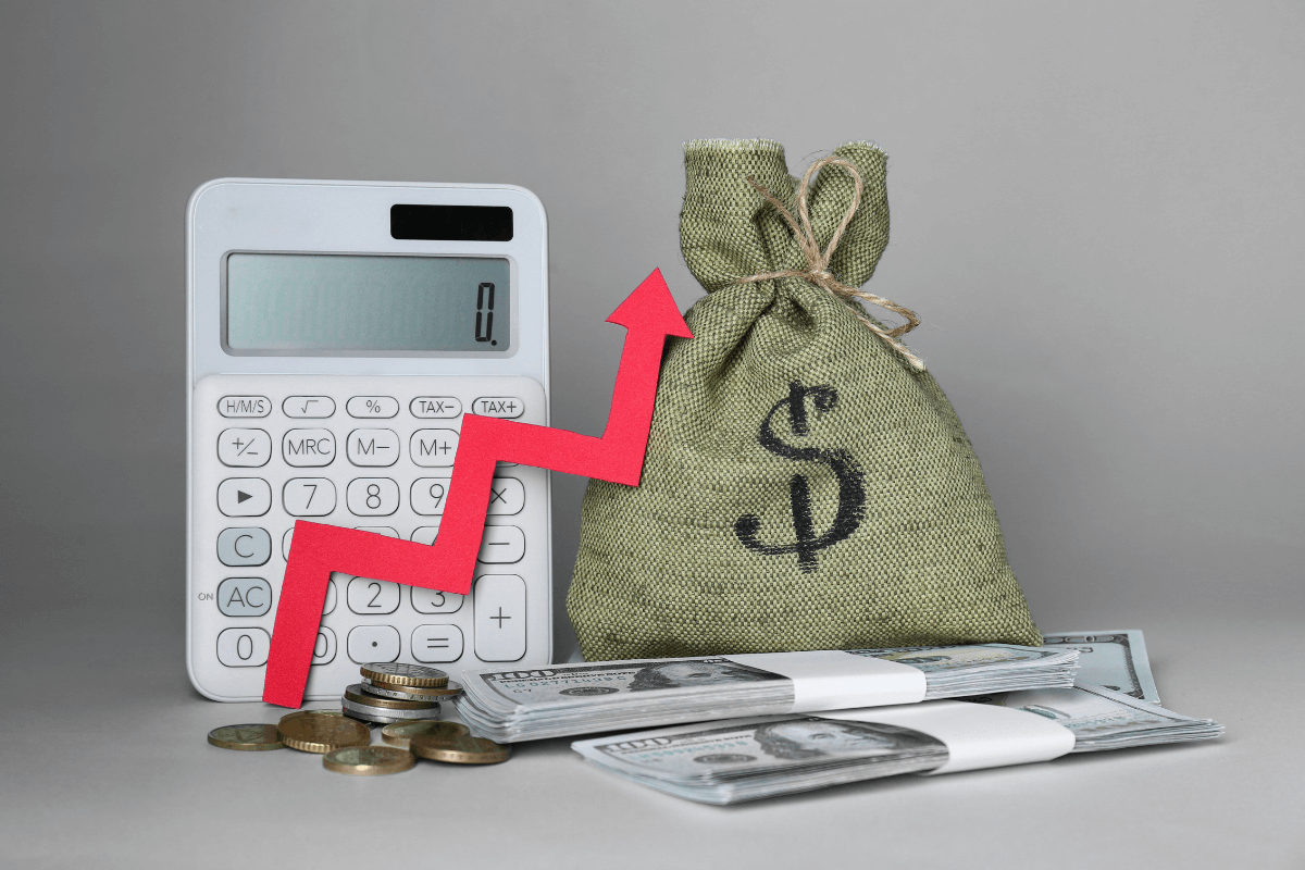 Calculadora branca ao lado de um saco de dinheiro e notas de dólar, com uma seta vermelha apontando para cima, simbolizando o aumento progressivo dos lucros.