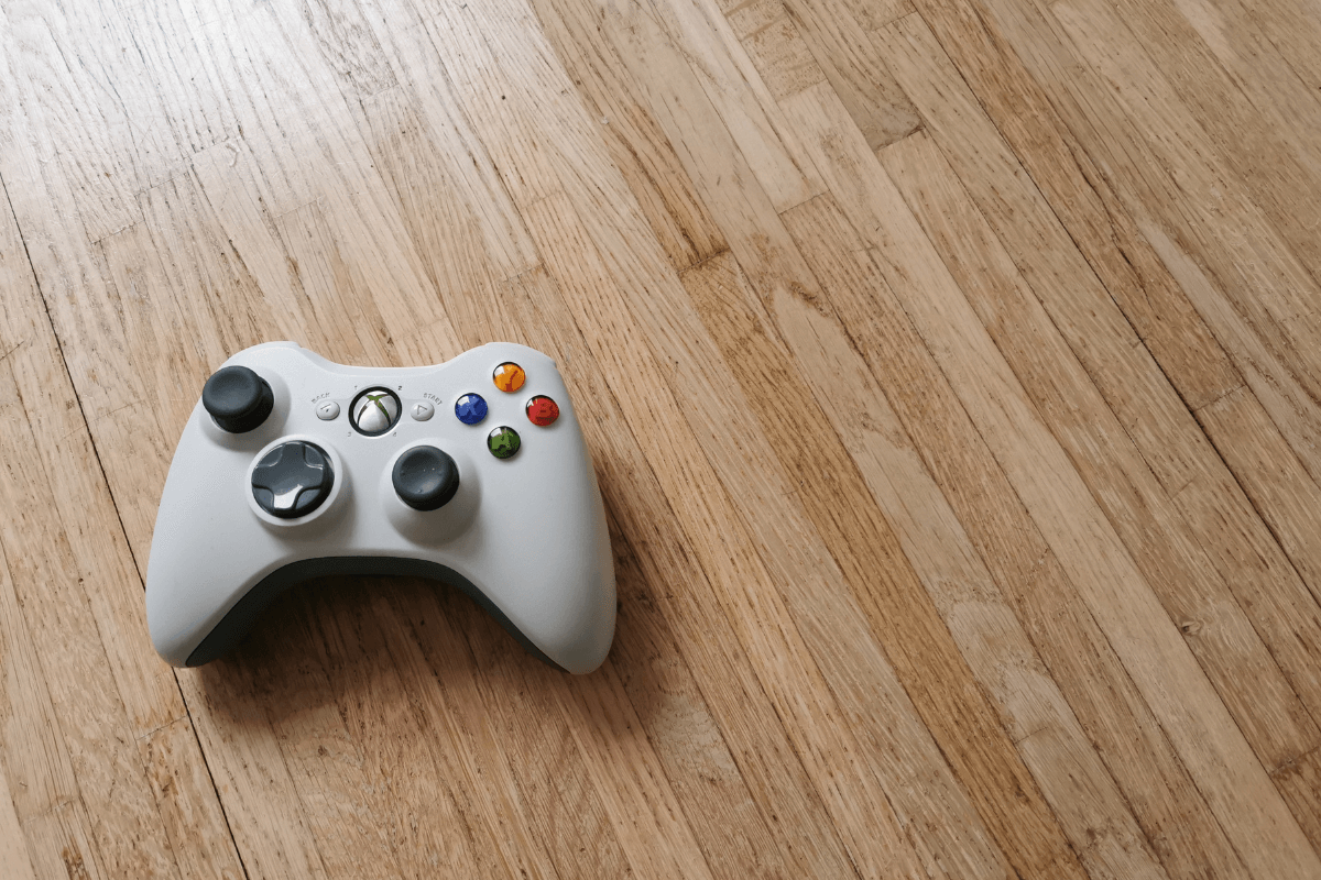 Controle de videogame Xbox branco sobre piso de madeira, ilustrando como o uso de jogos e missões ajuda a atingir a meta mensal do Microsoft Rewards.