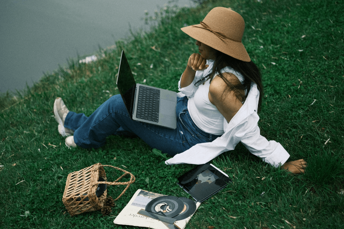 Mulher usando chapéu sentada na grama trabalhando em um notebook com um tablet ao lado.