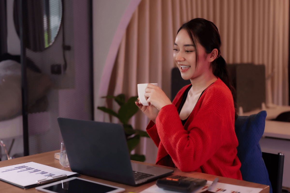 Mulher sorrindo sentada à mesa trabalhando no notebook com uma xícara de café na mão.