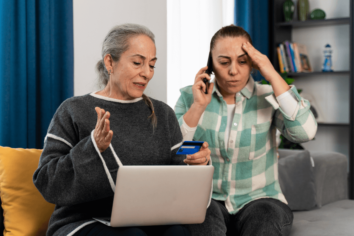 Senhora segurando um cartão de crédito com expressão de dúvida em frente ao notebook, enquanto outra mulher ao lado fala ao telefone parecendo tensa.