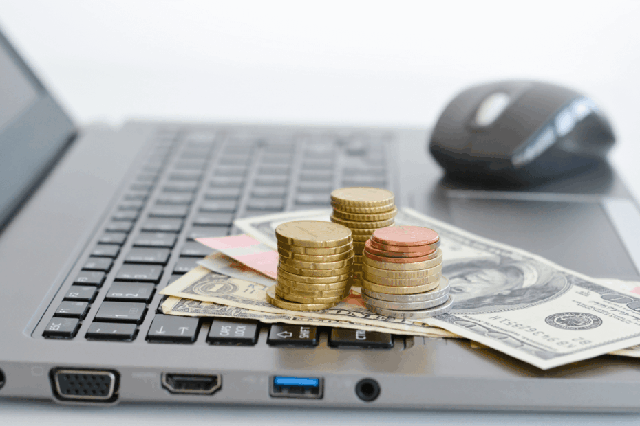 Pilha de moedas douradas e notas de dinheiro sobre o teclado de um notebook cinza, com um mouse ao fundo, ilustrando a monetização através de plataformas de pesquisa de mercado.