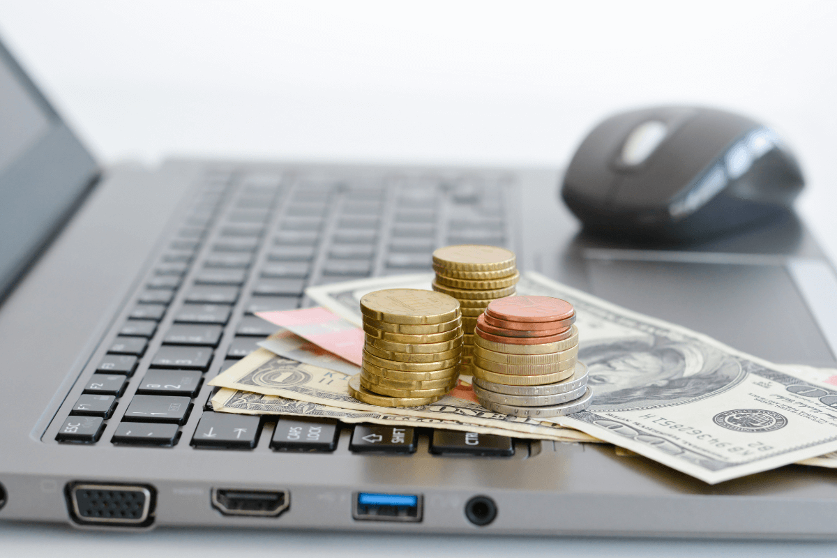 Pilha de moedas douradas e notas de dinheiro sobre o teclado de um notebook cinza, com um mouse ao fundo, ilustrando a monetização através de plataformas de pesquisa de mercado.