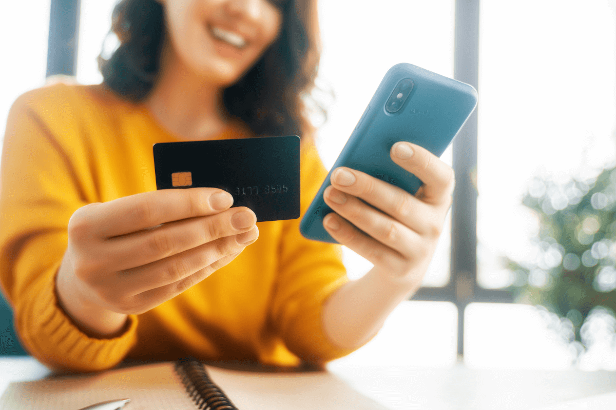 Mulher sorrindo com cartão de crédito e celular na mão, representando a facilidade de compra e o gatilho emocional que gera vendas imediatas na internet.