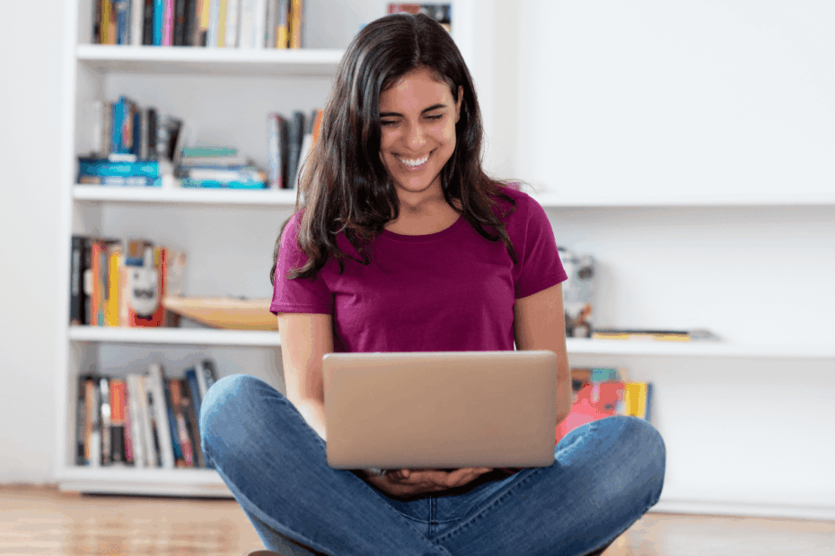 Mulher sorrindo sentada no chão em frente a uma estante de livros, usando um notebook no colo para acessar o site mais confiável para ganhar dinheiro respondendo pesquisas em casa.