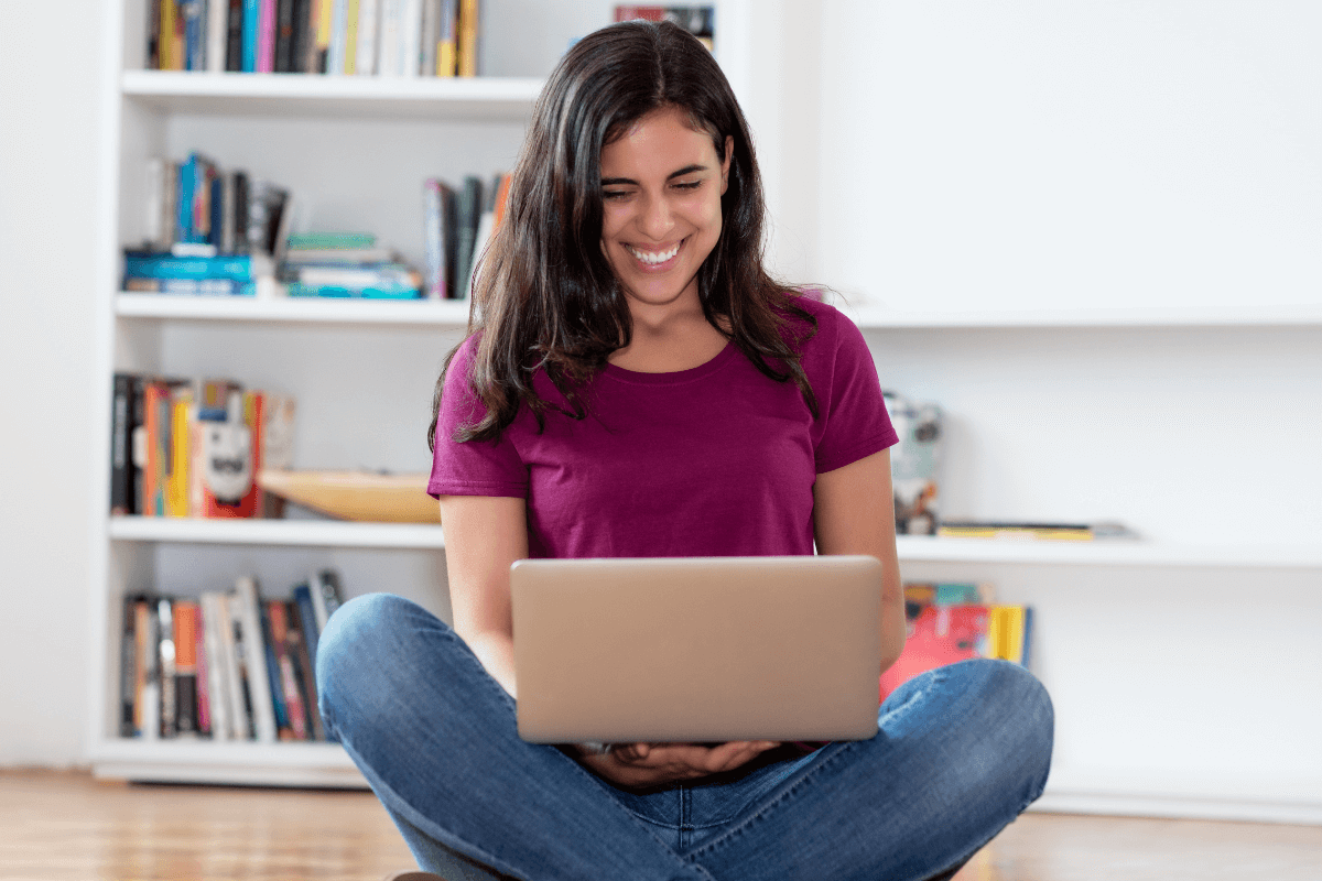 Mulher sorrindo sentada no chão em frente a uma estante de livros, usando um notebook no colo para acessar o site mais confiável para ganhar dinheiro respondendo pesquisas em casa.