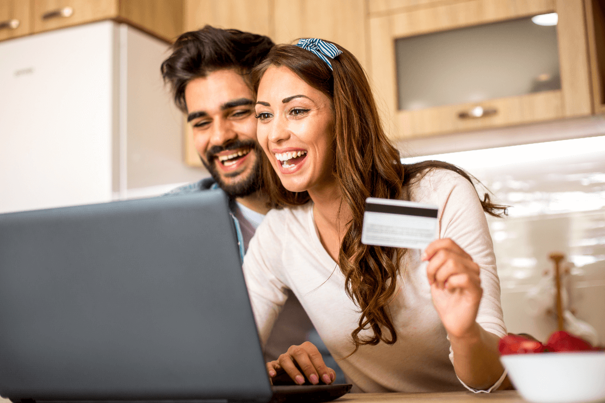 Casal sorrindo fazendo compras online com cartão de crédito e notebook na cozinha, ilustrando o perigo de afiliados usarem o próprio link para vendas familiares no mesmo endereço.